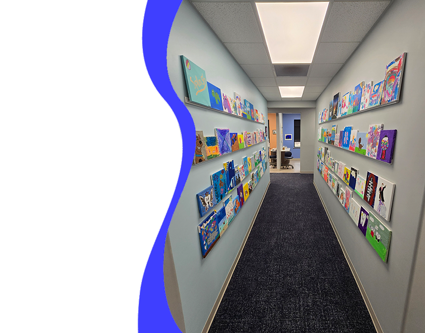 The image shows an interior view of a well-lit room with a blue and white color scheme. It appears to be a waiting area, possibly within a medical office, with shelves stocked with various items such as books, cards, and small gifts. The flooring is carpeted in neutral tones, and the walls are painted in a light hue. There s a large window on the right side of the room, allowing natural light to enter.